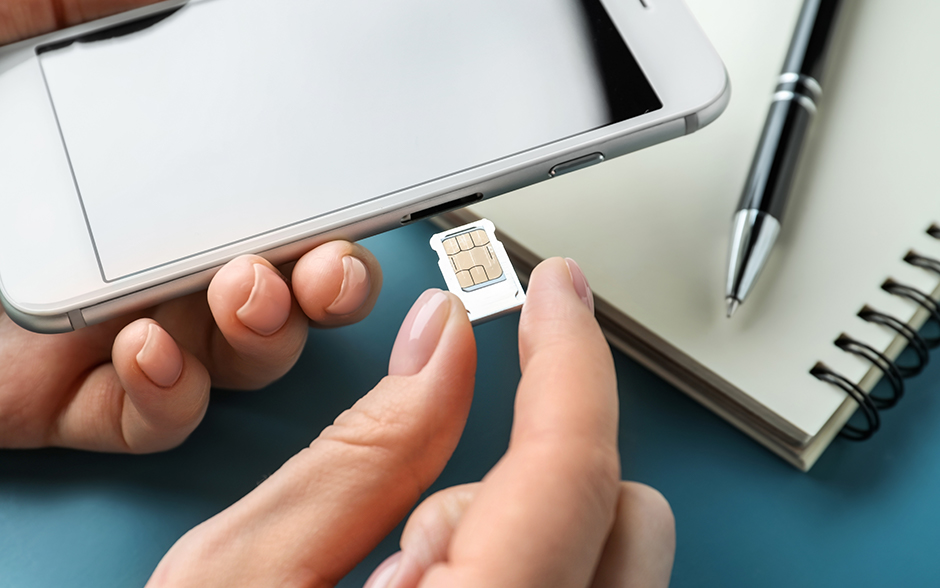 Close-up of hands inserting a SIM card into a smartphone, with a notebook and pen in the background on a blue surface, emphasizing a tech or communication setup.