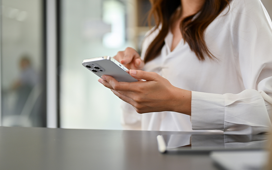 Professional woman in a white blouse using a smartphone at a modern office desk, symbolizing connectivity and workplace productivity.