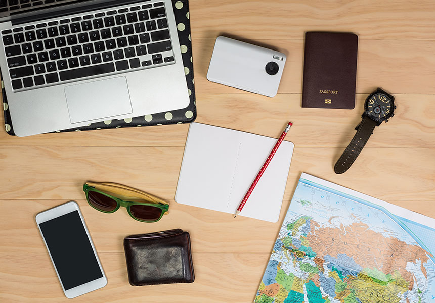 Flat lay photo of a map, wallet, smartphone, sunglasses, notebook, pencil, laptop, camera, passport, and watch on table.