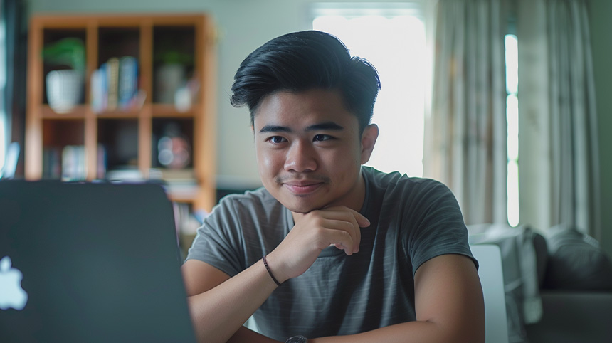 Young Asian man in his home office holding his chin thoughtfully while looking at his laptop.