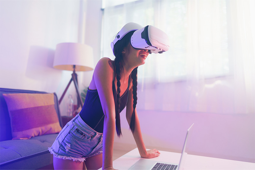 Young Asian woman wearing VR goggles at home, smiling widely as if enjoying what she's seeing.