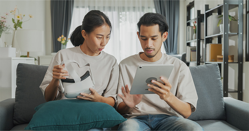 Young Asian couple sitting on a couch, holding VR glasses and a digital tablet, ready to enjoy a virtual reality experience.