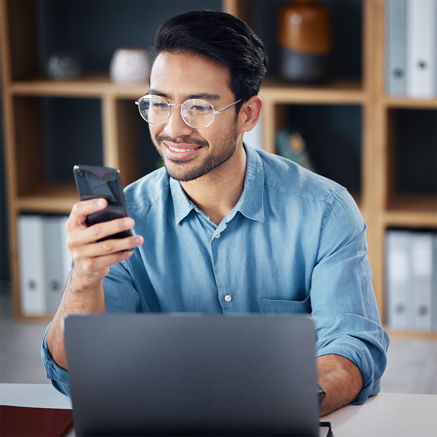 Young Asian man in front of his laptop, looking at his phone.