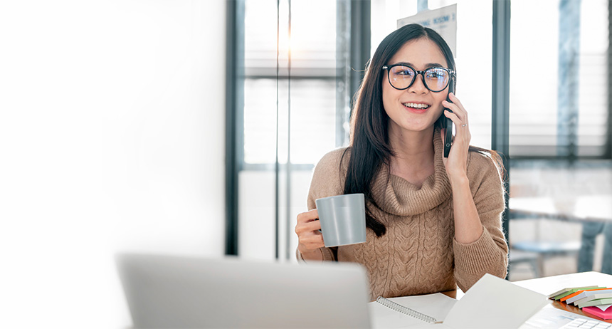 Cheerful woman sitting at a desk in a bright home office, talking on her smartphone while gesturing, with a tablet, pen, and documents in front of her.