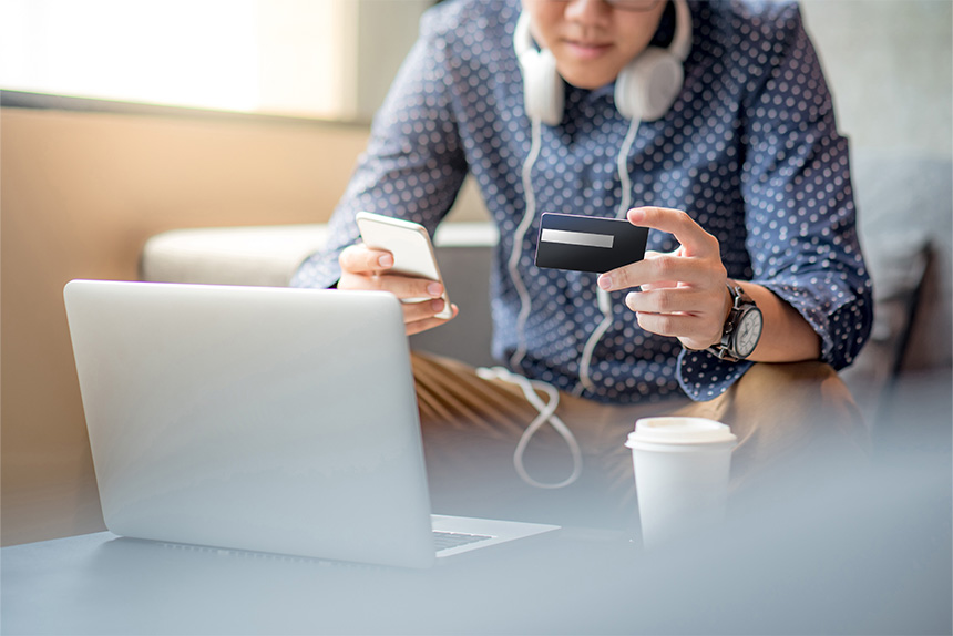 Smiling man holding a smartphone and a credit card, preparing to make an online payment at a bright office desk.