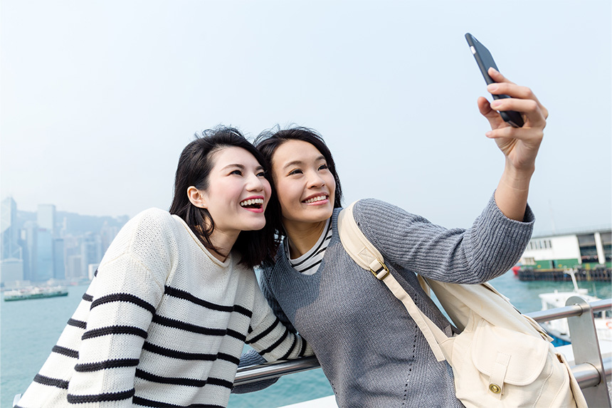 Two young girls take a selfie on the deck of a ferry ship with a view of a city in the background.