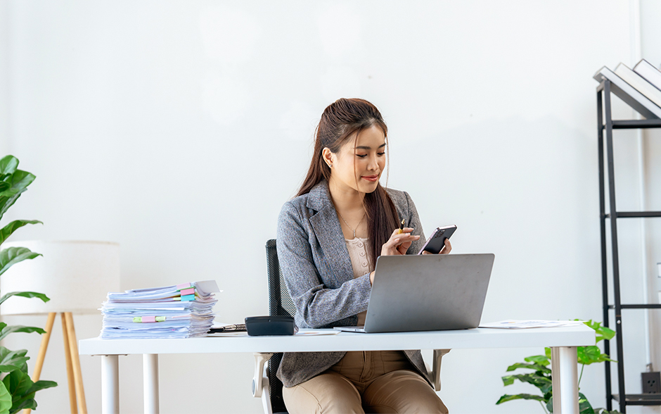 A professional woman sitting at a desk, using her smartphone while working on a laptop, with stacks of documents nearby