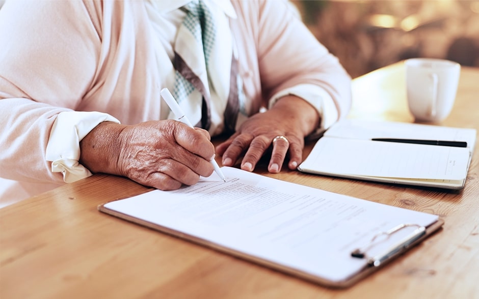 An elderly woman carefully fills out a form on a clipboard at a wooden table