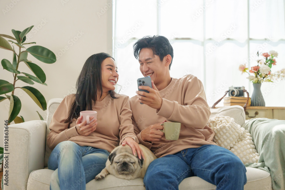 Two women share a joyful moment on a sofa in a naturally lit, cozy room, one holding a mug while they look at a smartphone.