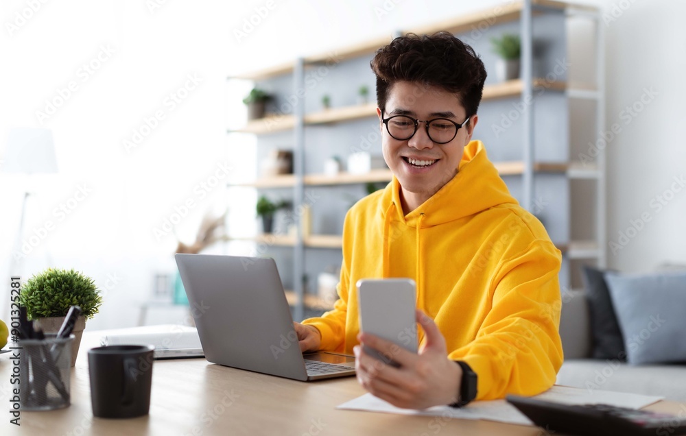 Person in a yellow hoodie using a smartphone and laptop at a desk in a bright room.