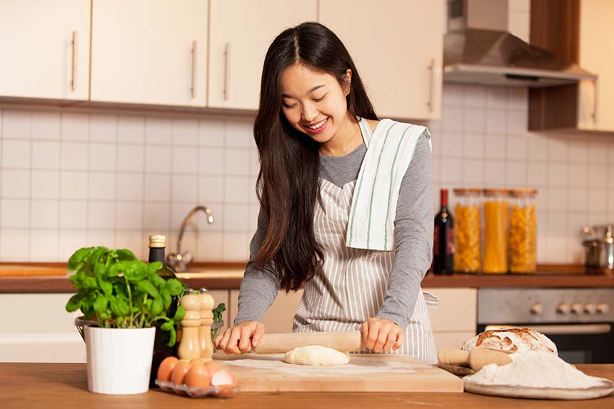 A woman in a bright kitchen rolling out dough with a smile, surrounded by fresh bread, eggs, and herbs, embodying a cozy baking scene.