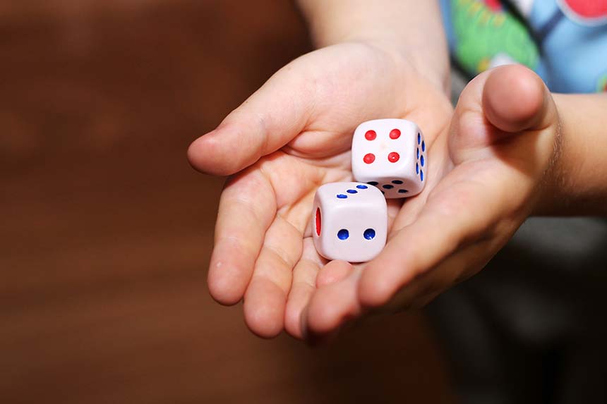 A pair of hands holds two white dice with red and blue dots, set against a warmly lit, blurred background.
