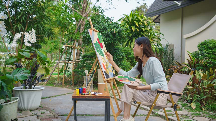 A woman seated outdoors, painting a vibrant canvas on an easel, surrounded by lush greenery and garden tools.