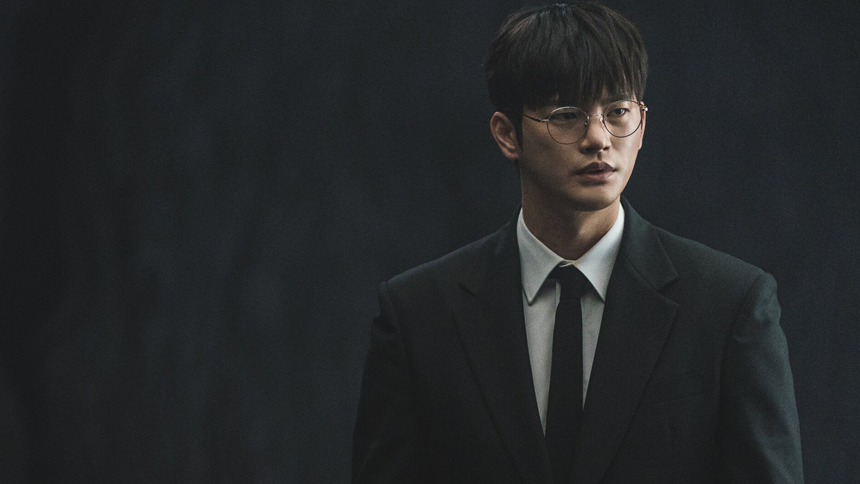 A young man in a black suit and tie stands against a dark, moody background, his serious expression highlighted by soft, dramatic lighting.