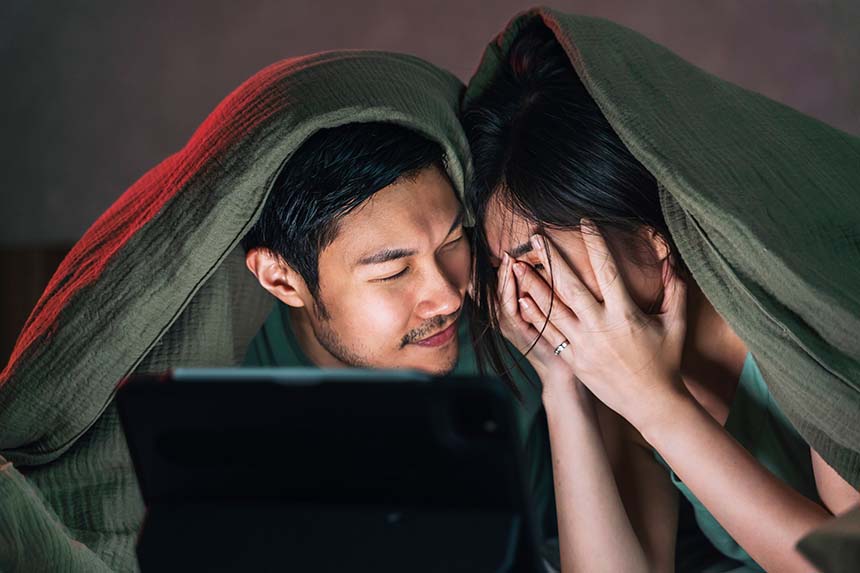 A couple snuggles under a blanket in a dimly lit room, watching something on a screen, with the woman playfully covering her face.