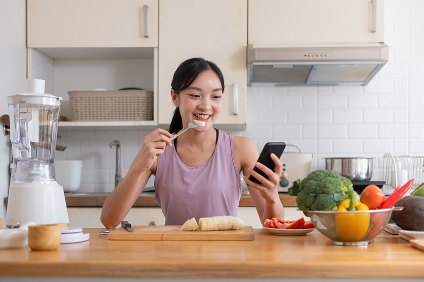 A woman enjoys a healthy snack while browsing her phone in a bright kitchen, surrounded by fresh vegetables and a blender.