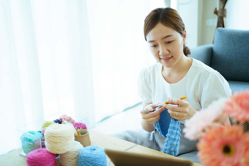 Two hands cradling soft white yarn balls, with a green plant and a knitted sweater adding a cozy touch in the background.
