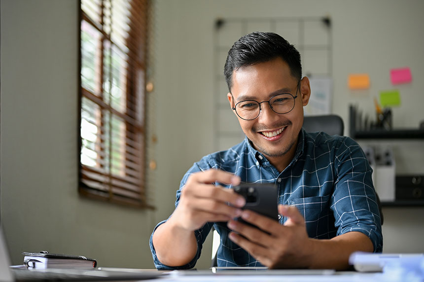 Young Asian man looking at his smartphone while seated on his desk.