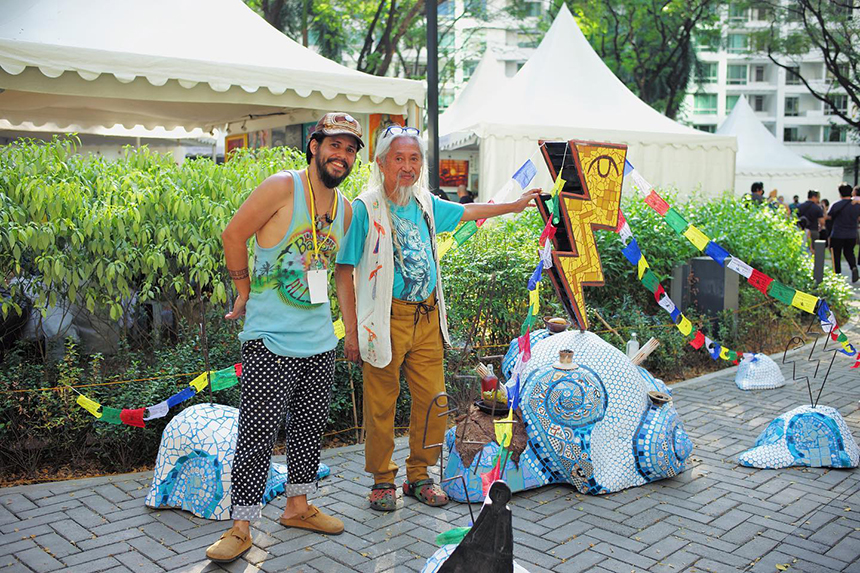 Two men pose happily at an outdoor art festival, surrounded by colorful sculptures, flags, and artistic displays, with tents and greenery in the background.