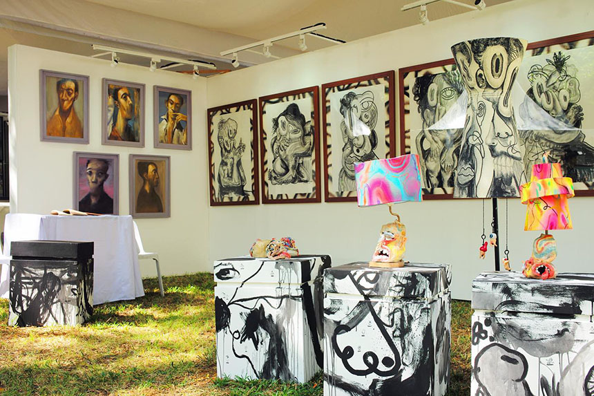 An eclectic outdoor art display features surreal portraits on the walls, abstract sculptures, and colorful lamps atop painted pedestals, creating a vibrant and imaginative atmosphere.