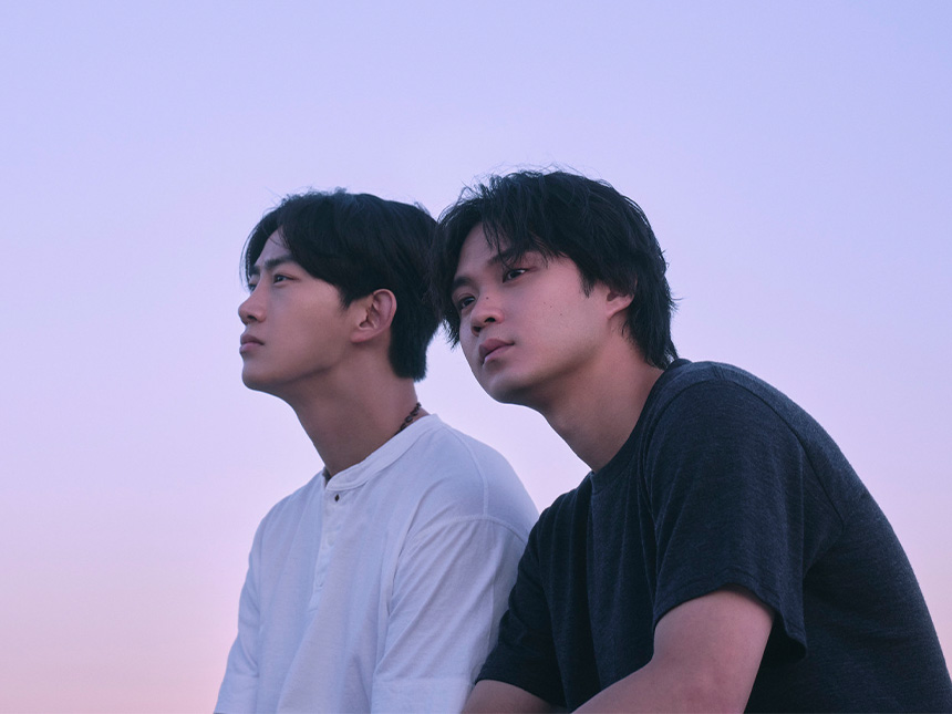 Two young men sit together gazing into the distance, with a soft purple sky at dusk as the background.