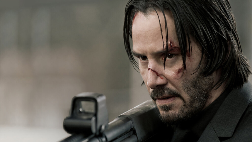 Close-up of a wounded man with long hair and a beard, holding a weapon and looking intensely forward.