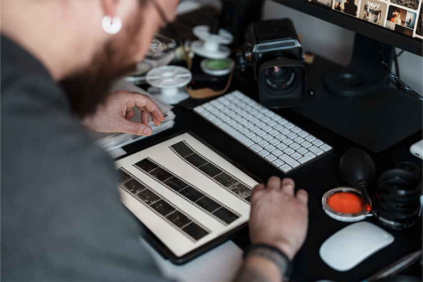 Person examining film strips on a lightbox at a cluttered desk with camera equipment and a computer monitor displaying thumbnails.
