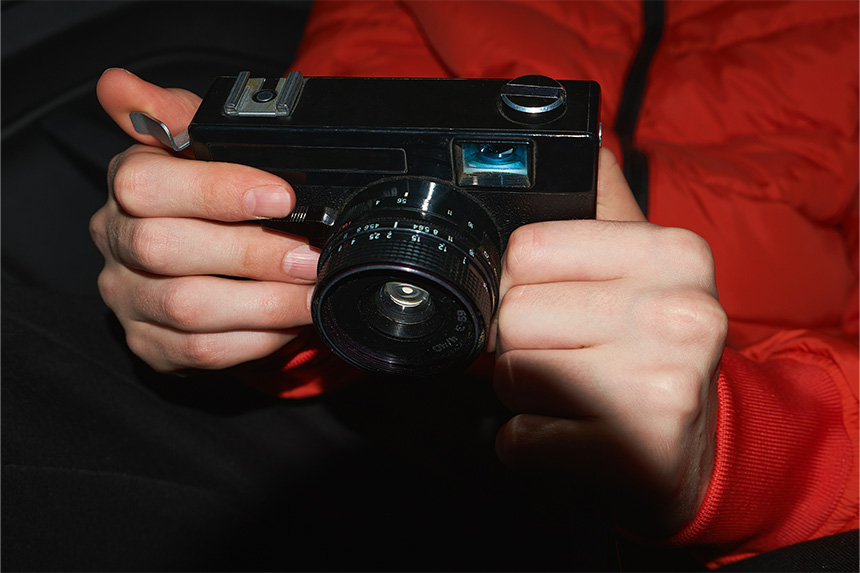 Close-up of hands holding a vintage black camera with a red jacket in the background.