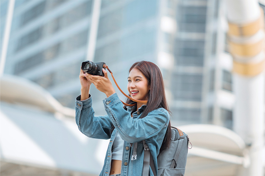 A young woman in a denim jacket, smiling while holding a camera, with city buildings in the background.
