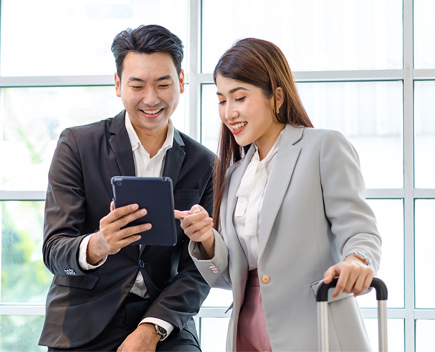A man in a dark suit and a woman in a gray blazer look at a tablet together, smiling.
