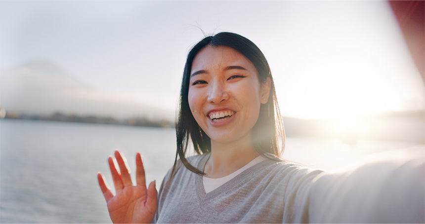 A woman smiling and taking a selfie near a body of water with a mountain in the background, during sunset or sunrise.