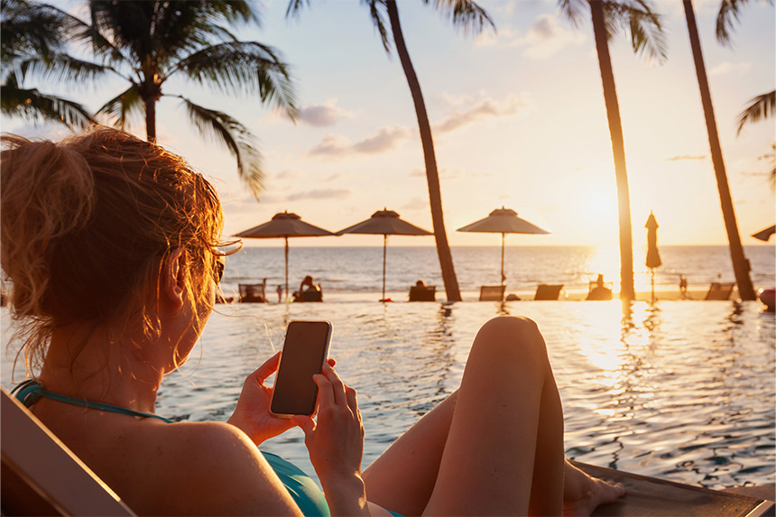 Person by an infinity pool at sunset, holding a smartphone. Palm trees and beach chairs in the background.