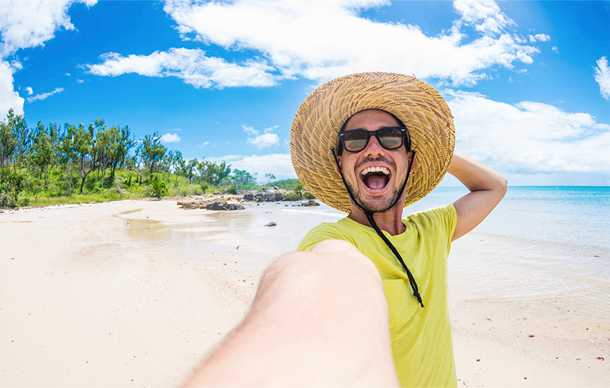 Person taking a selfie on a sunny beach wearing a straw hat and sunglasses
