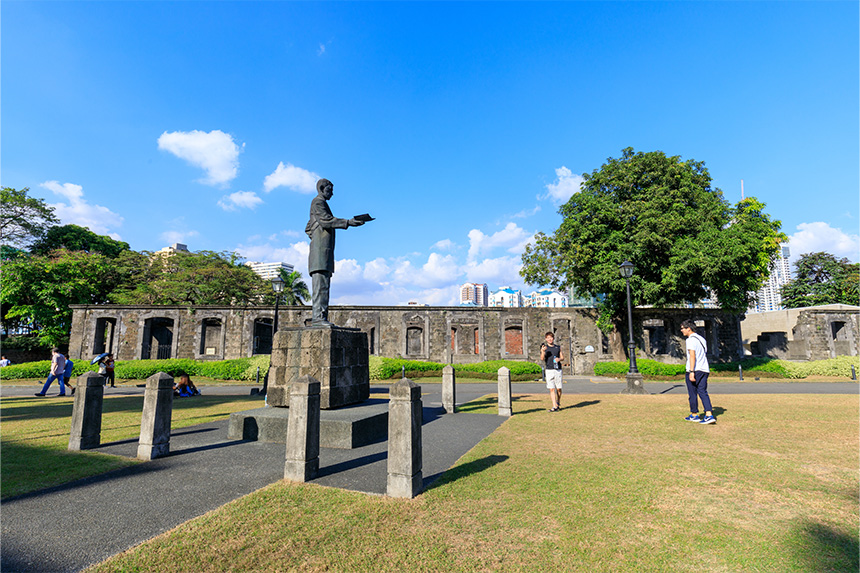 A statue of a man in a park with historical stone buildings and modern cityscape in the background.