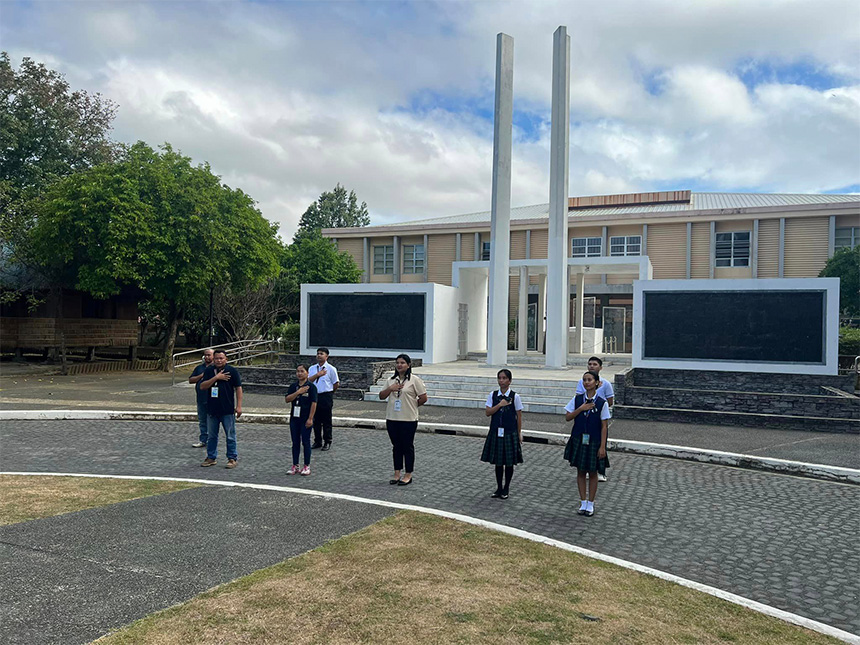 A group of people standing outside near a monument with tall columns and black plaques in front of a beige building.
