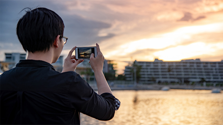 Person taking a photo of a sunset near a waterfront with buildings in the background.