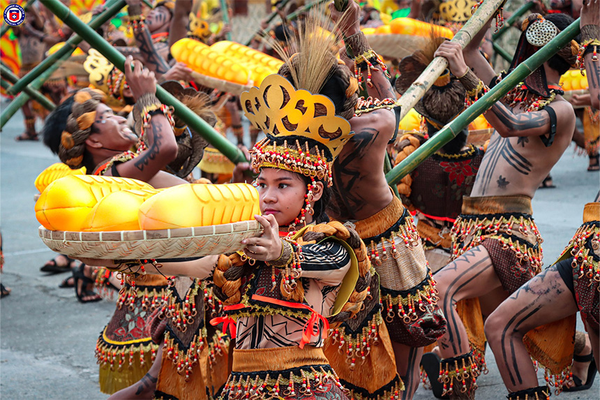 Dancers in traditional attire holding bamboo poles, with one in front carrying a basket of yellow fruits.