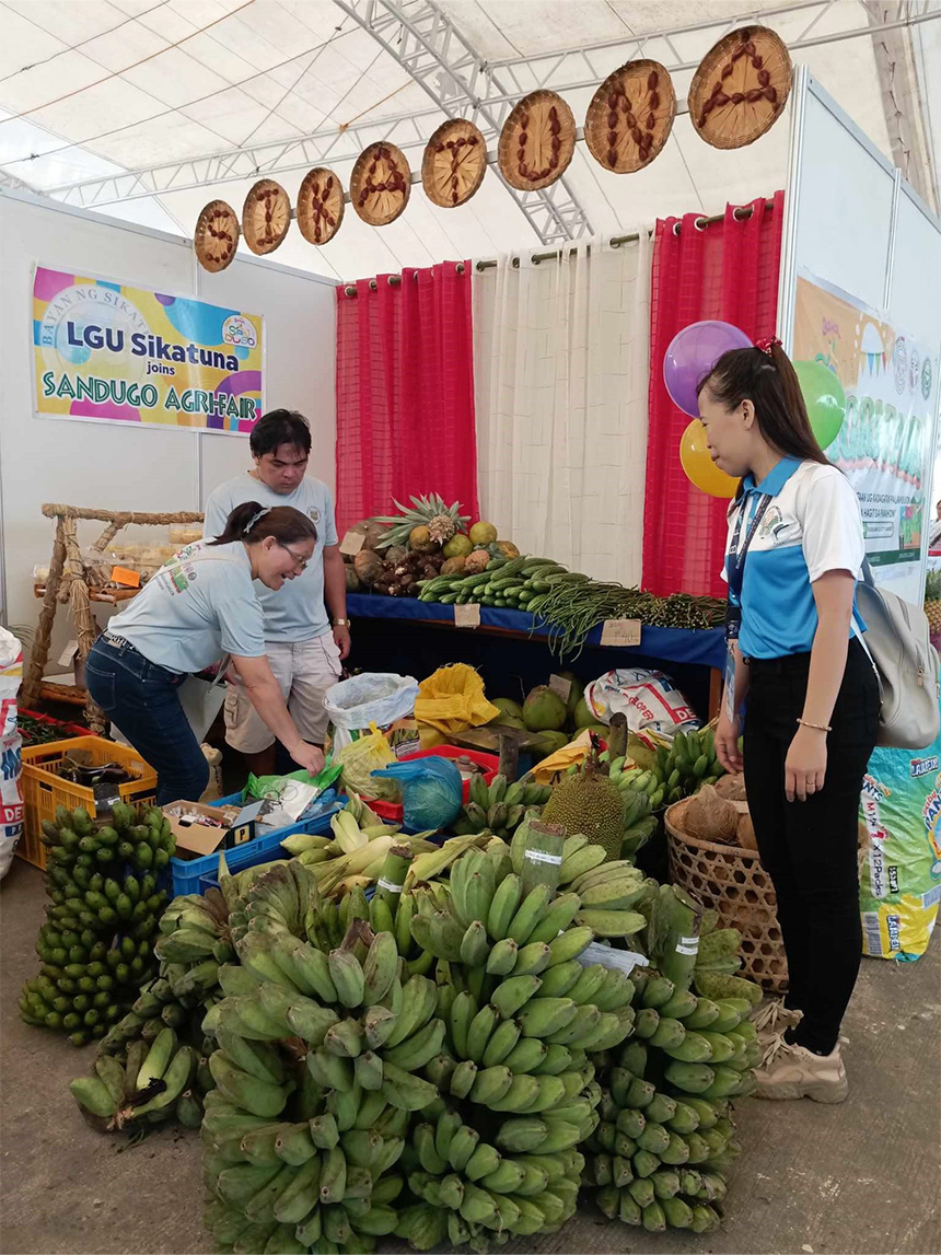 Market stall with fruits and vegetables under a tent with "LGU Sikatuna joins SANDUGO AGRIFAIR" sign.