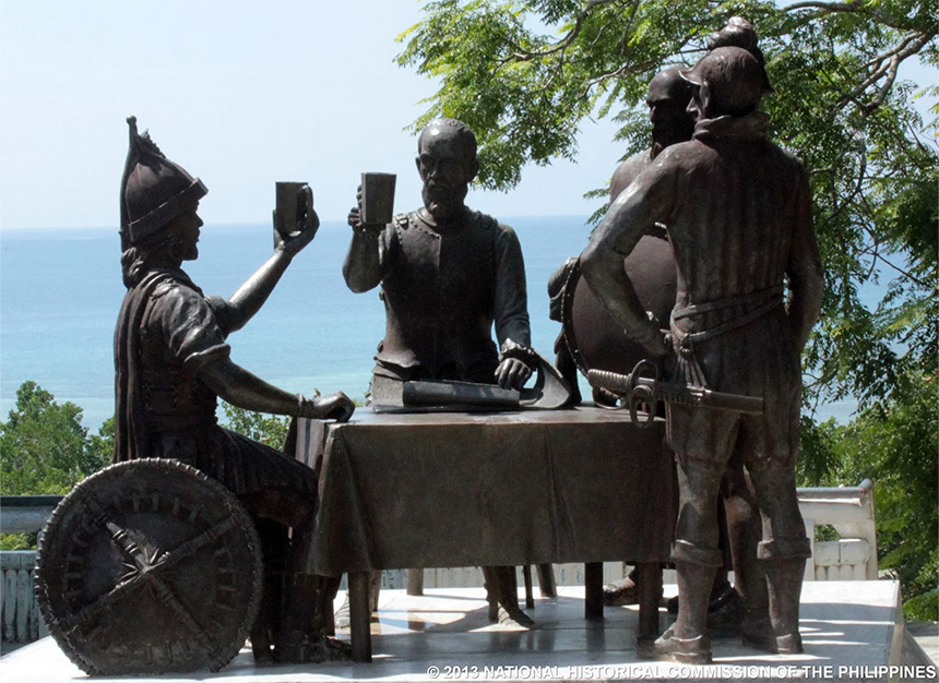 Bronze statue of historical figures around a table, with ocean in the background.
