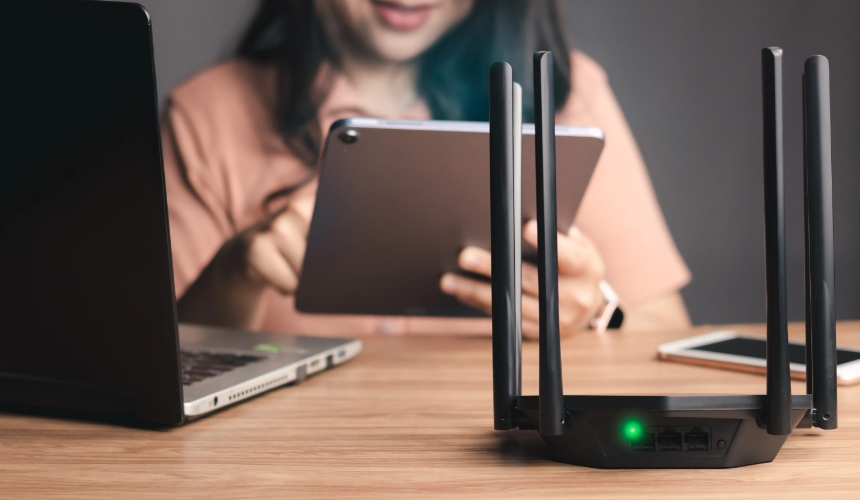 A black router with four antennas on a table, with a person using a tablet in the background.