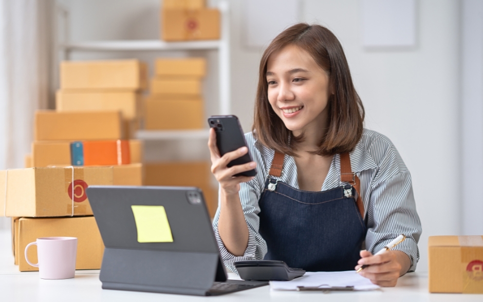An Asian businesswoman, wearing a striped shirt and apron, smiles while looking at her smartphone, surrounded by boxes of orders.