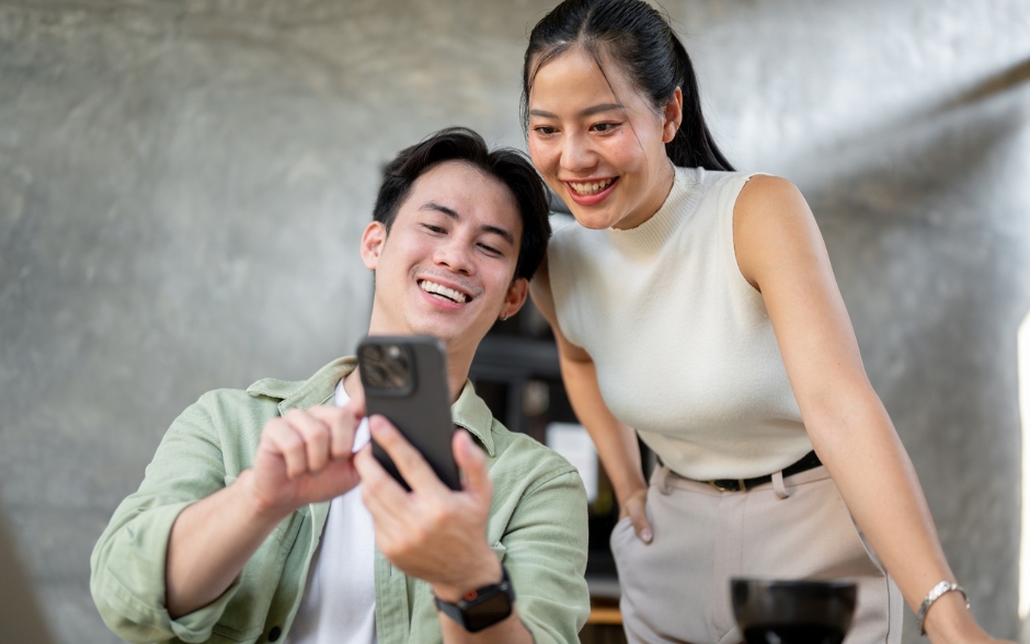 A smiling young couple looking at a smartphone together in a casual setting, conveying connection and shared enjoyment.