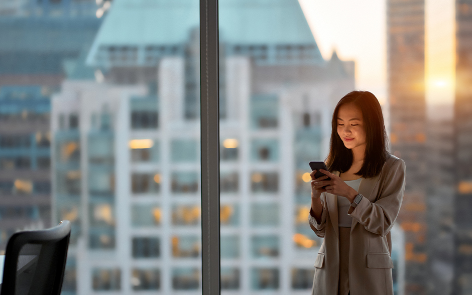 Smiling woman using a smartphone in a modern office with a cityscape view at sunset.