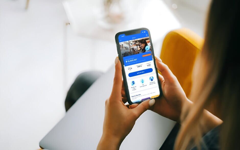 A person holding a smartphone with a screen showing GGives page, seated on a yellow chair indoors.