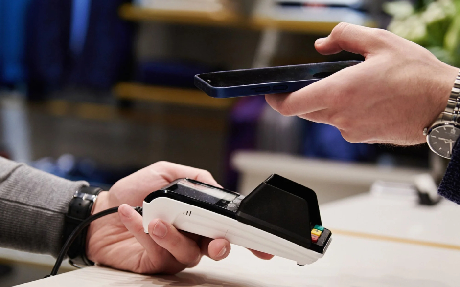 A person holds a smartphone above a portable card reader held by another person across a counter.