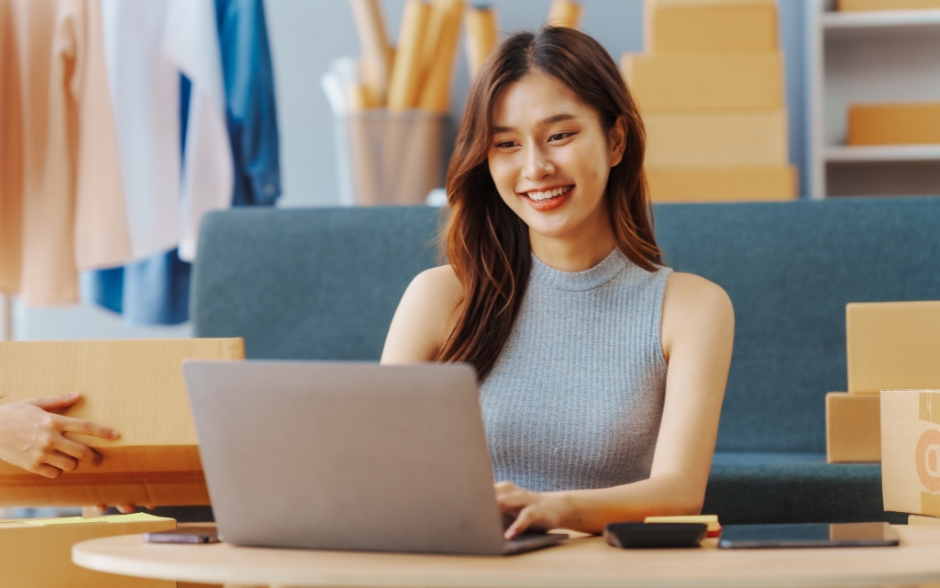 A woman with a smile sitting at a table, working on a laptop indoors.