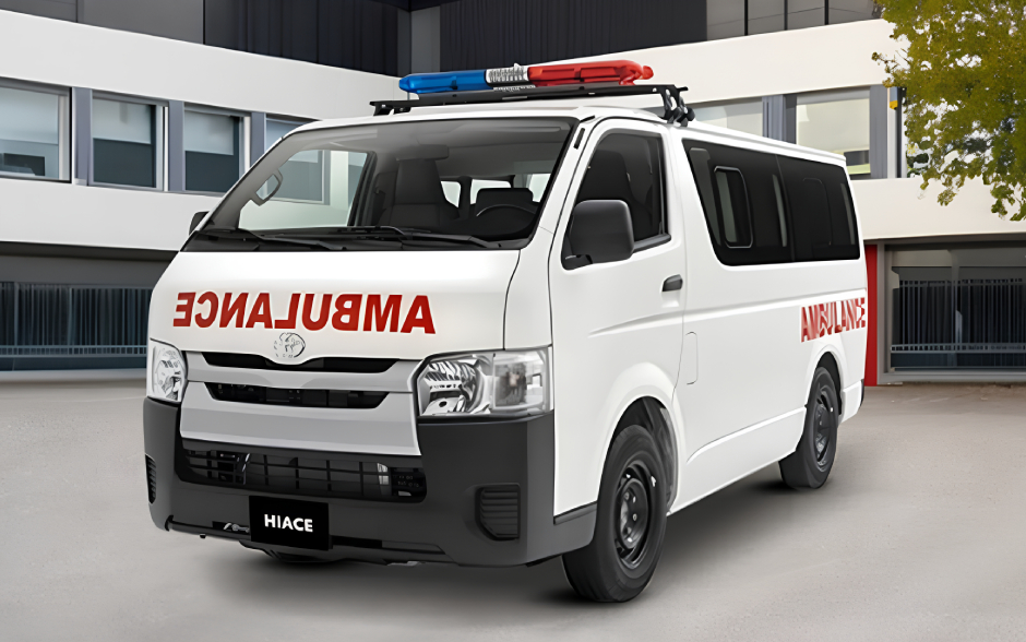 A white ambulance van with "AMBULANCE" written in reverse on the front, parked near a modern building.