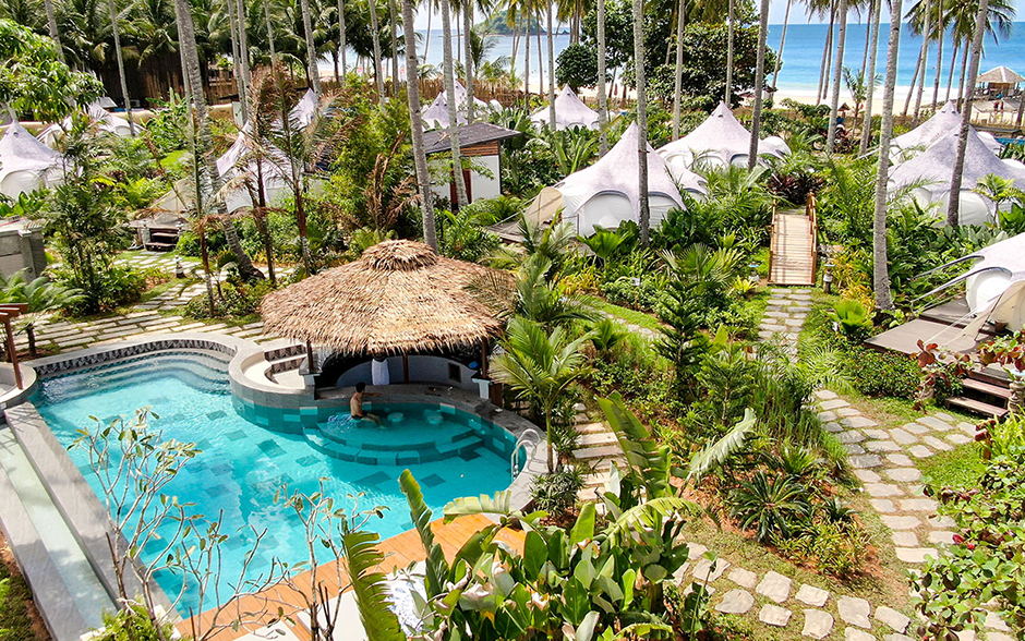 A photo of white glamping tents with navy blue details, set amidst lush greenery and palm trees on the beach in El Nido, Palawan, Philippines.