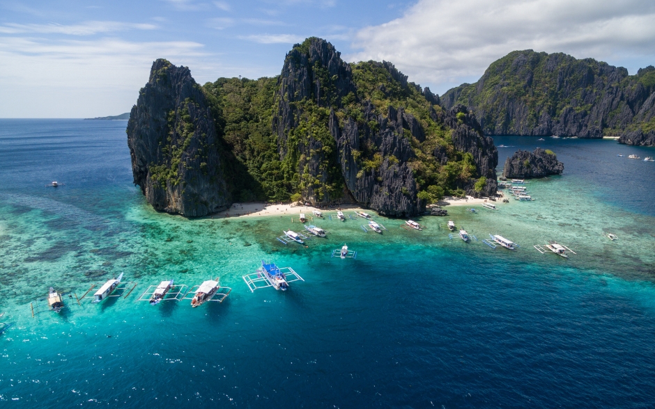 Aerial view of the beautiful scenery in El Nido, with towering cliffs and boats on the clear blue water, surrounded by a white sand beach on the island of Palawan.