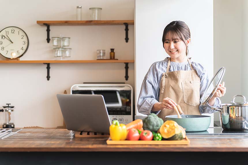 A woman in a modern kitchen wearing an apron, stirring a pot on the stove while referencing a laptop, with colorful vegetables displayed on the countertop.